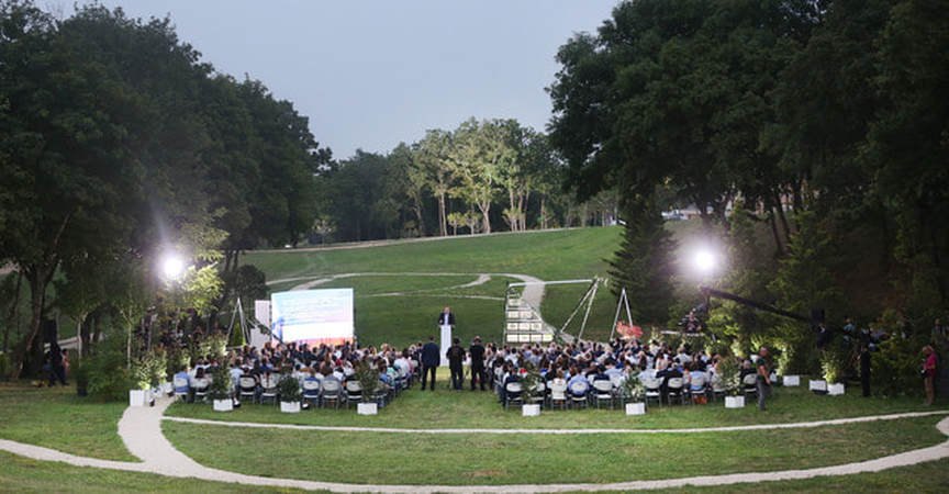 An open-air evening conference session on a lawn, with an audience seated before a lit presentation screen